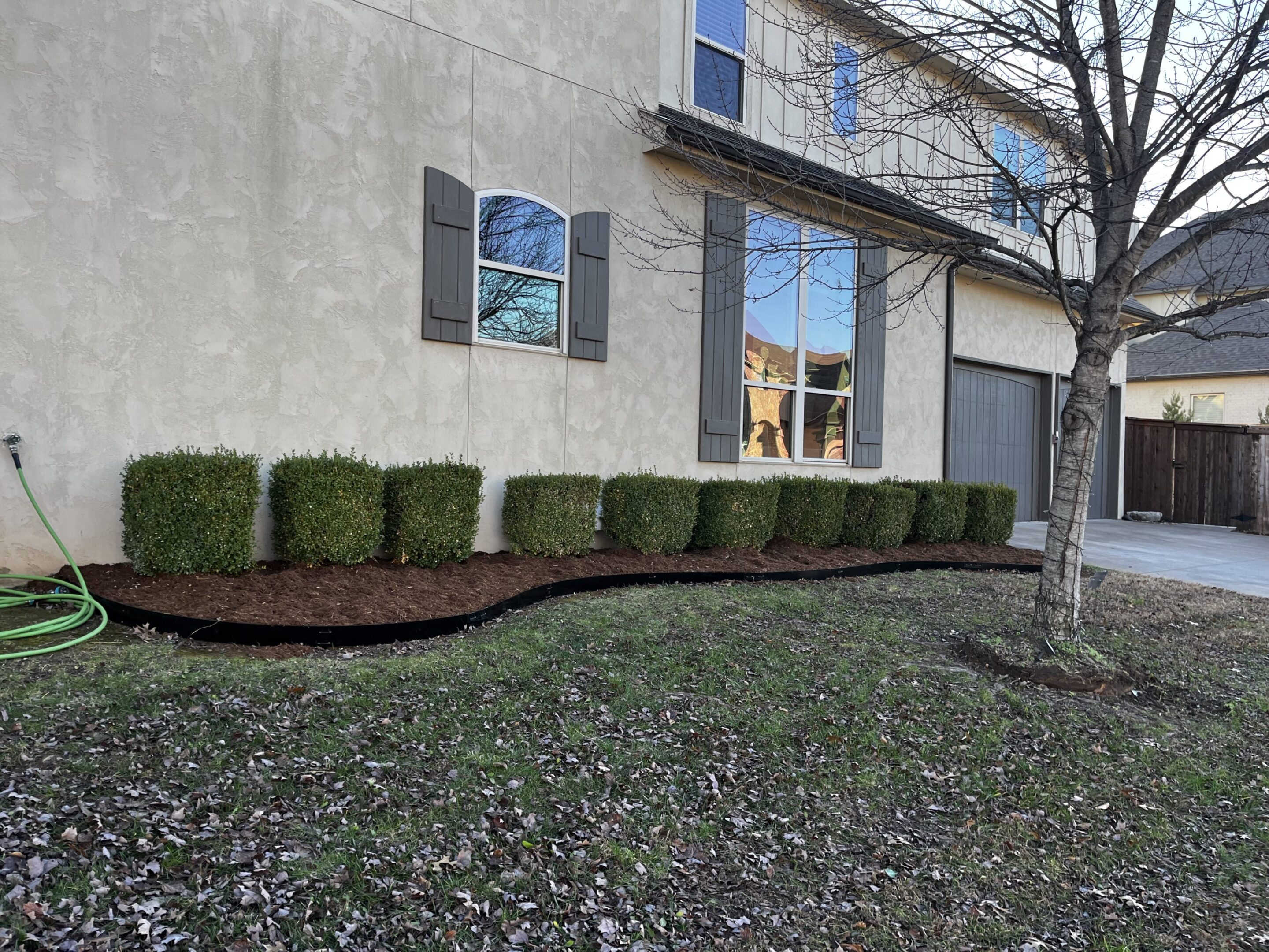 Neatly trimmed bushes line the side of a house with a window and a garage door.